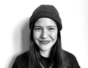 Eine lächelnde Person in einem schwarzen Shirt und Mütze vor einem weißen Hintergrund, in Schwarz-Weiß fotografiert.