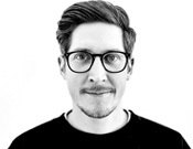 Ein Mann mit Brille und Bart schaut in die Kamera, schwarz-weiß Foto, kurzer Haarschnitt und dunkles Oberteil.