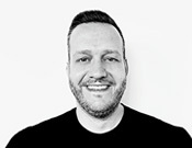 Porträt eines lächelnden Mannes mit Bart in Schwarz-Weiß. Er trägt ein dunkles T-Shirt und hat kurzes, dunkles Haar.