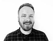 Ein lächelnder Mann mit Bart und kariertem Hemd vor weißem Hintergrund in Schwarzweiß.