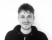 Ein lächelnder Mann mit kurzem Haar trägt einen dunklen Kapuzenpullover vor einem weißen Hintergrund.