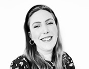 Schwarz-Weiß-Foto einer lachenden Person mit langem Haar, Stirnband und Ohrring, in blumigem Oberteil vor weißem Hintergrund.