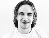 Schwarz-weiß Porträt eines lächelnden Menschen mit schulterlangem Haar und T-Shirt vor weißem Hintergrund.