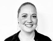 Schwarz-weiß-Porträt einer lächelnden Person mit hochgestecktem Haar, großen Ohrringen und einer Halskette vor weißem Hintergrund.