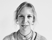 Schwarz-Weiß-Porträt einer Person mit langem Haar, einem Pullover und mehreren Halsketten, die in die Kamera lächelt.