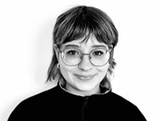 Person mit Brille und schulterlangem Haar lächelt leicht in die Kamera. Sie trägt einen dunklen Pullover vor weißem Hintergrund.