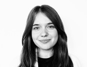 Porträt einer lächelnden Person mit langen Haaren vor einem weißen Hintergrund, in Schwarz-Weiß aufgenommen.