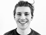 Junger Mann mit welligem Haar lächelt in die Kamera. Schwarz-weiß Porträt vor weißem Hintergrund.