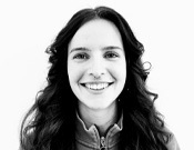 Junge Frau mit langen lockigen Haaren lächelt in die Kamera. Schwarz-weiß Porträt vor hellem Hintergrund.