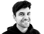 Ein lächelnder Mann mit dunklen Haaren und Bart, trägt einen schwarzen Kapuzenpulli vor weißem Hintergrund.