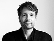 Ein Mann mit Bart und kurzen Haaren blickt freundlich in die Kamera. Er trägt ein dunkles Jackett vor hellem Hintergrund.