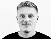 Schwarz-weiß Porträt eines lächelnden Mannes mit hellem Haar und Bart, in einem dunklen Shirt vor weißem Hintergrund.