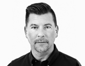 Porträt eines Mannes mit dunklem, zurückgekämmtem Haar und Bart, in einem dunklen Hemd. Der Hintergrund ist weiß.