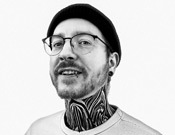 Mann mit Brille, Bart und Mütze lächelt. Sein Hals ist auffällig tätowiert. Schwarz-weiß Foto auf weißem Hintergrund.