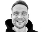Ein lächelnder Mann trägt einen Hoodie, vor einem weißen Hintergrund. Sein Haar ist kurz geschnitten und er schaut direkt in die Kamera.