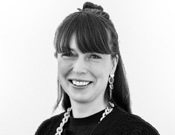 Frau mit langen Haaren, einer Kette und Ohrringen, lächelnd vor neutralem Hintergrund, in Schwarz-Weiß aufgenommen.