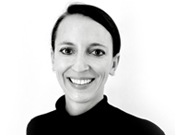 Porträt einer lächelnden Person mit dunklem Haar, das nach hinten gebunden ist. Sie trägt einen dunklen Rollkragenpullover vor weißem Hintergrund.