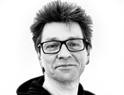 Schwarz-weiß-Foto einer Person mit Brille und hochstehendem Haar, die leicht lächelt. Sie trägt einen dunklen Pullover.