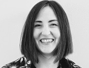 Eine lächelnde Person mit schulterlangem Haar, in Schwarz-Weiß aufgenommen. Sie trägt eine gemusterte Oberbekleidung.