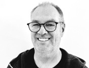 Ein lächelnder Mann mit Brille und Bart in Schwarz-Weiß. Er trägt eine dunkle Jacke vor einem hellen Hintergrund.