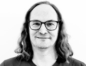 Porträt eines lächelnden Menschen mit langen, glatten Haaren und Brille vor einem weißen Hintergrund in Schwarz-Weiß gehalten.