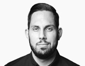 Ein Mann mit Bart, dunklem Haar und schwarzem Hemd blickt ernst in die Kamera. Der Hintergrund ist weiß.