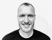 Porträt eines lächelnden Mannes mit kurzen Haaren und schwarzem Oberteil vor weißem Hintergrund, in Schwarz-Weiß.