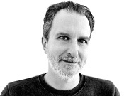 Ein Mann mit kurzem Haar, hellem Bart und neutralem Gesichtsausdruck blickt in die Kamera vor einem weißen Hintergrund.