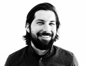 Mann mit Bart und schulterlangem Haar lächelt. Schwarzweiß-Foto vor hellem Hintergrund.
