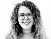 Frau mit lockigem Haar und Brille lächelt in die Kamera. Schwarz-weiß Porträt vor weißem Hintergrund.