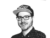 Ein Mann mit Brille und Bart trägt eine gemusterte Kappe und ein Hemd mit Punkten. Er lächelt in die Kamera. Schwarz-weiß-Bild.